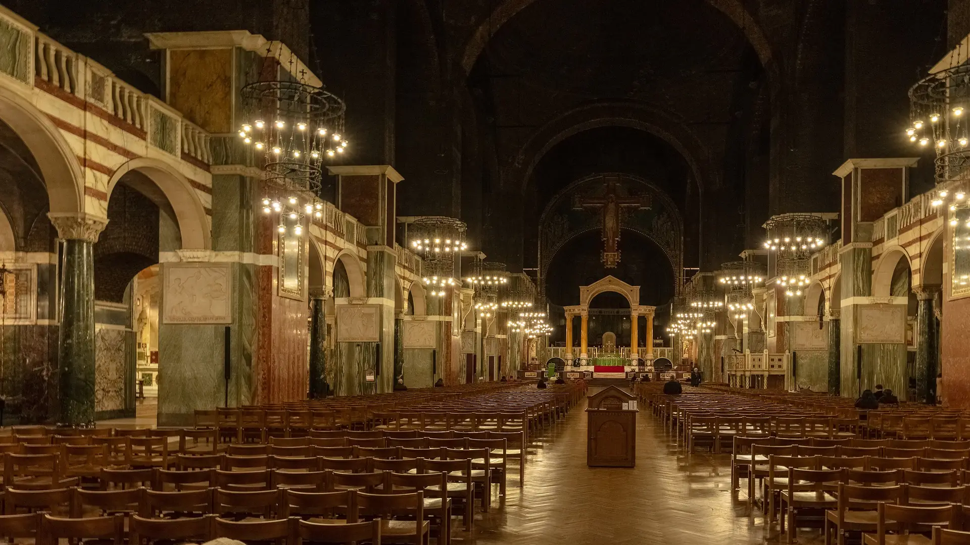 The nave of Westminster Cathedral at night, looking east past striped marble arcades and hanging lamps to the great Rood cross and the baldacchino over the high altar.